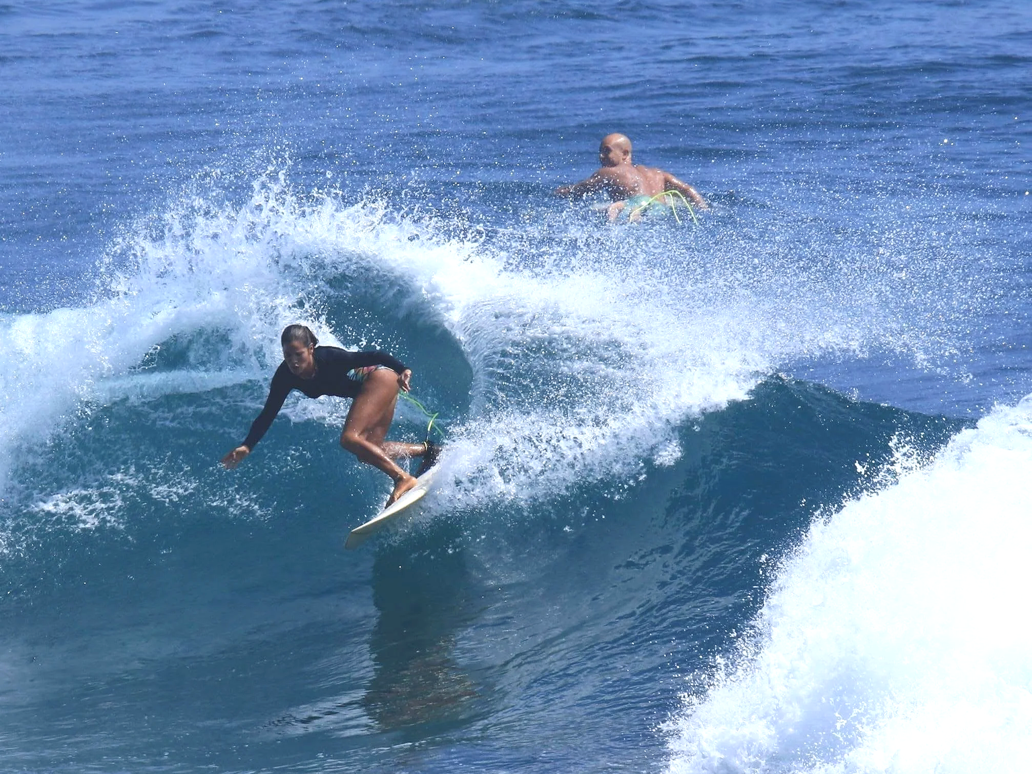 A woman surfing on a wave with a man in the background on a surfboard in the ocean.