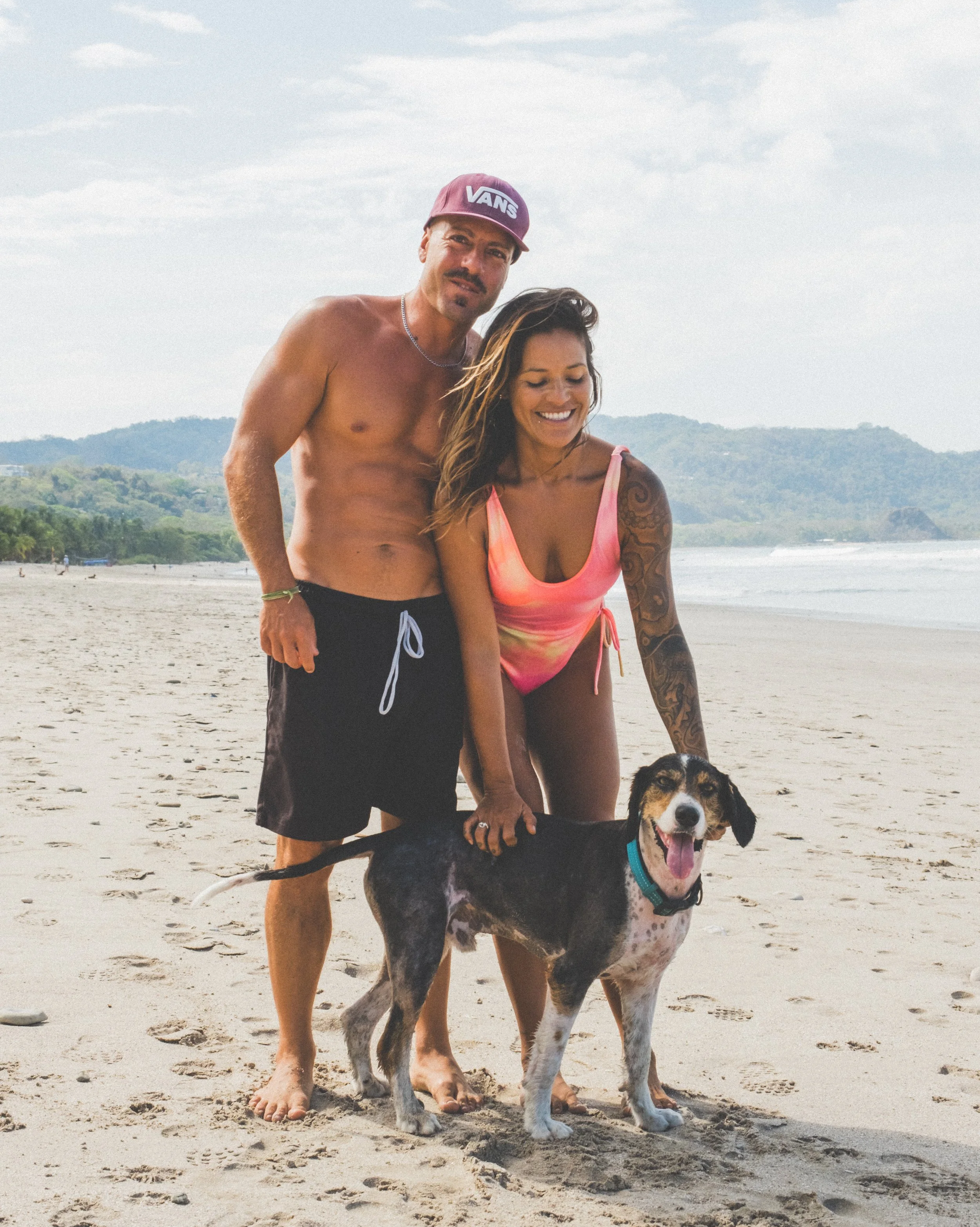 A man and a woman in beachwear pose with a happy dog on a sandy beach with hills and ocean in the background.