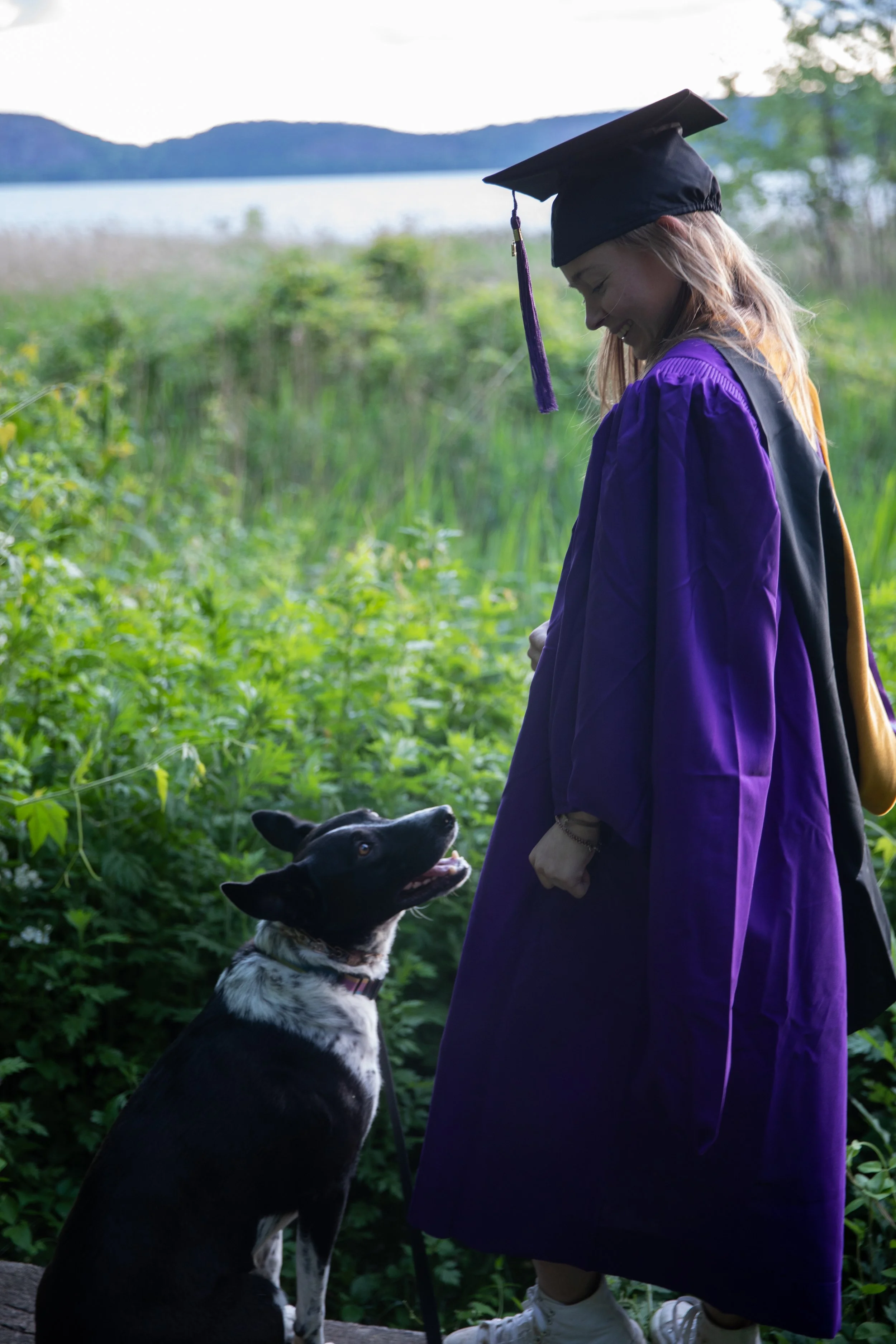 A young woman in a purple graduation gown and cap with a tassel stands outdoors near a lake and green bushes, smiling at her black and white dog that is sitting next to her and looking up.