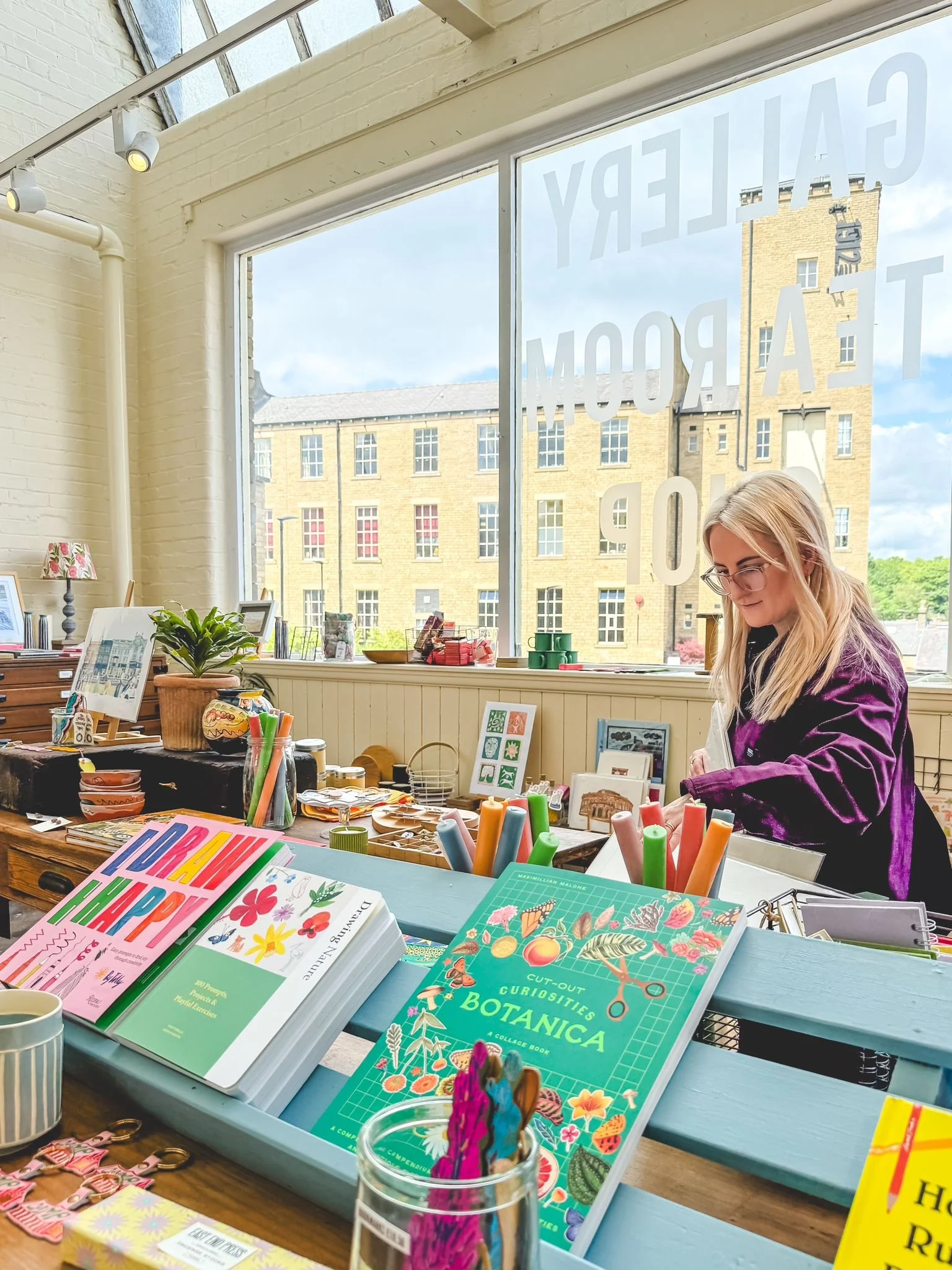 Inside a store with a woman arranging items on a table near a large window. The table features colorful books, stationery, and decorative objects. The window has large lettering and reveals a brick building outside.