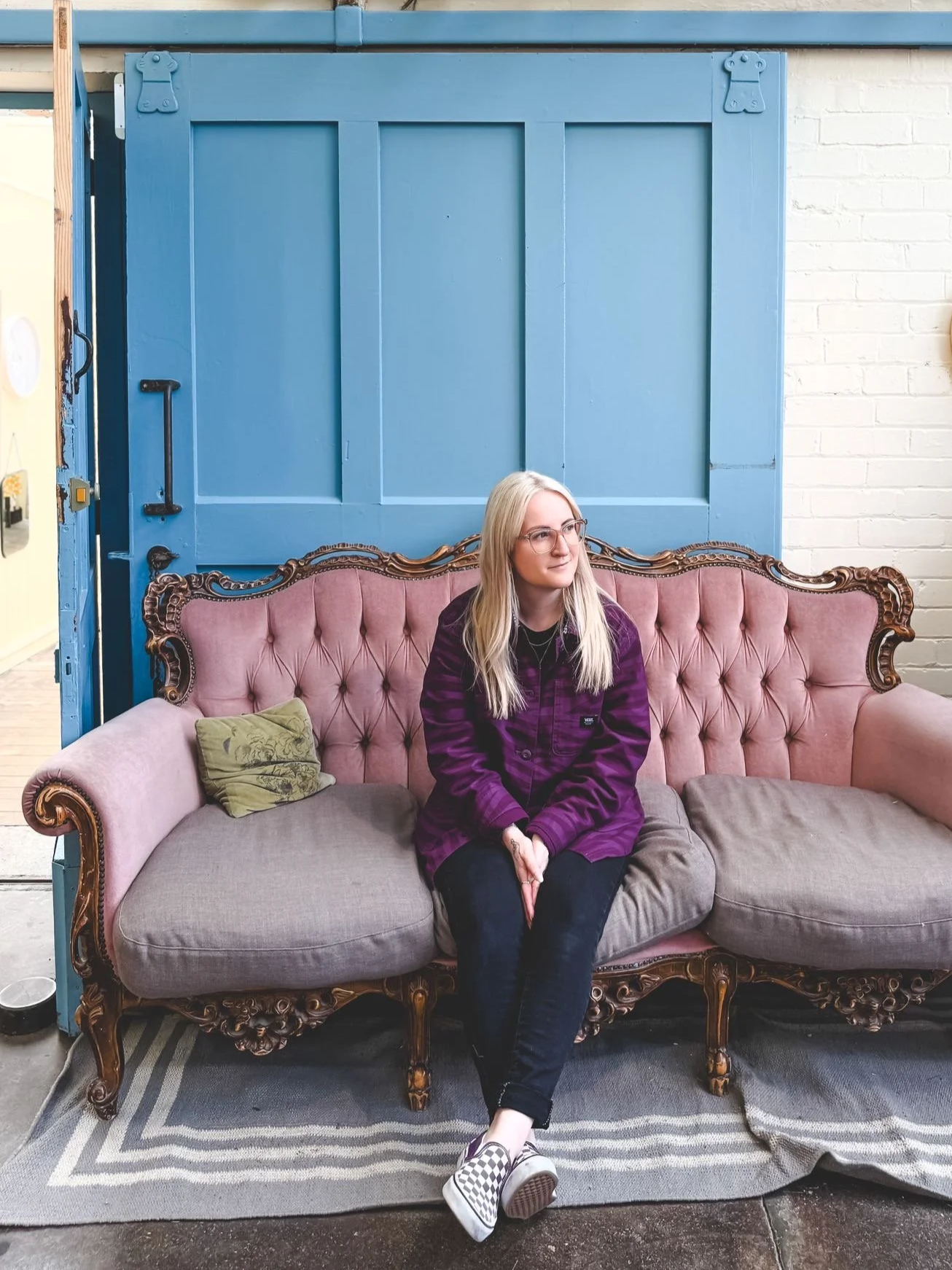 A young woman with blonde hair, glasses, and a purple plaid jacket sitting on a vintage pink tufted sofa with wooden accents, in front of a large blue sliding door with a black handle, next to a white brick wall.