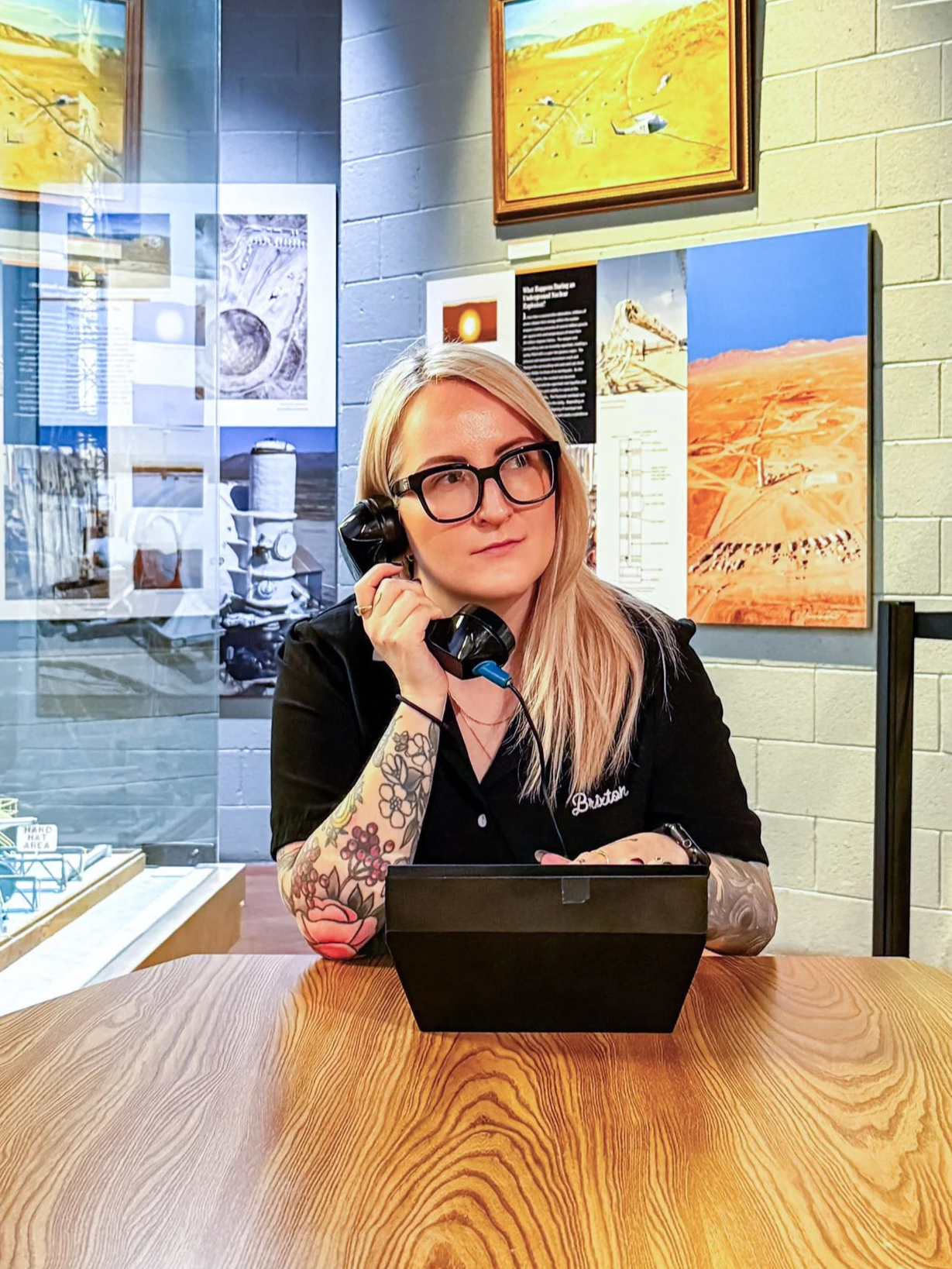 A woman with blonde hair, black glasses, and tattoos on her right arm, wearing a black shirt, is sitting at a wooden table with a black telephone receiver to her ear. She appears to be in an art gallery or museum, with colorful paintings and informational posters on the wall behind her.