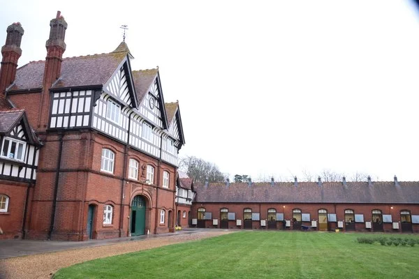 A large historic brick building with Tudor-style architecture and a green door, adjacent to a long red brick wall with small windows, and a well-maintained grassy lawn in front.