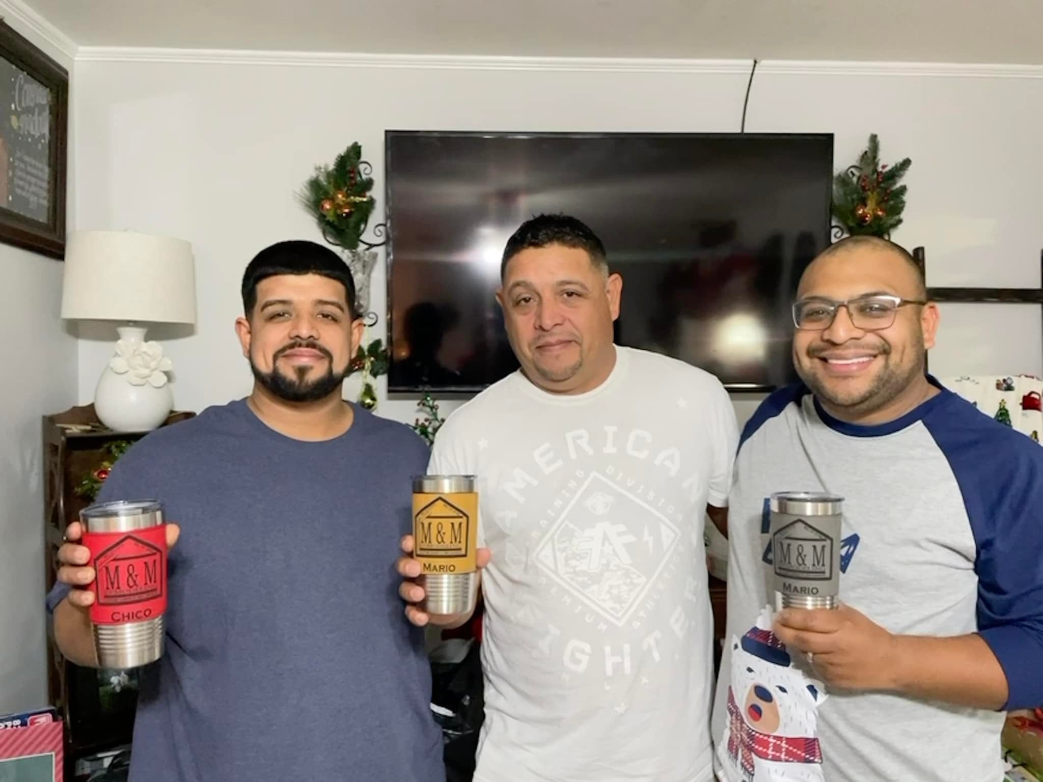 Three men standing indoors, holding personalized tumblers, smiling at the camera with a Christmas-themed background.