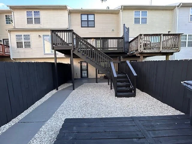 Backyard with a two-level wooden deck, black stairs, and black fencing, with a pebble-filled ground and neighboring houses in the background.