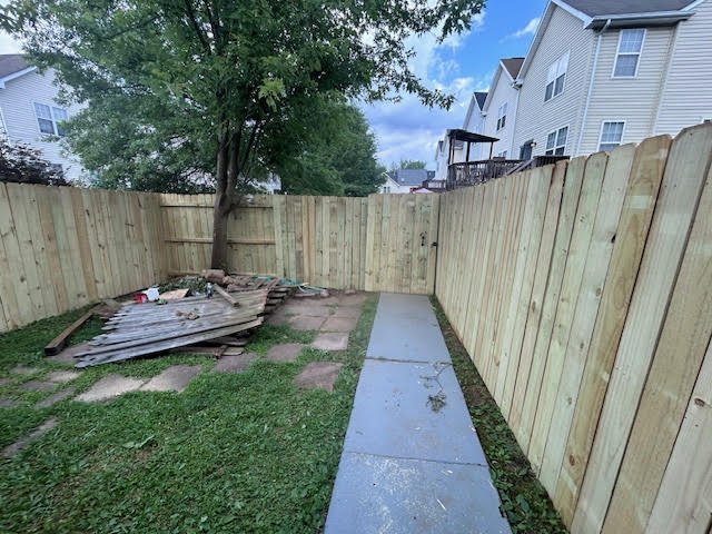 Backyard with a wooden fence, a concrete path, a pile of wooden debris, and trees with neighboring houses in the background.