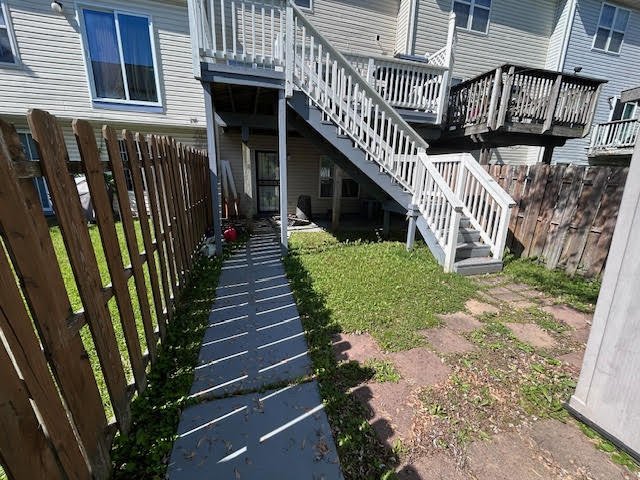 Backyard view showing a concrete pathway leading to a wooden staircase attached to an elevated deck of a house, with a wooden fence on the left and garden area on the right.