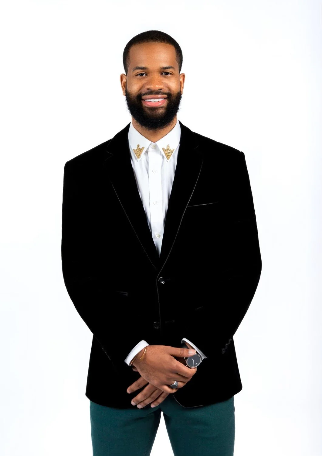 A man in a black blazer, white shirt with gold decorative pins on the collar, and teal pants, standing against a white background, smiling and looking at the camera.