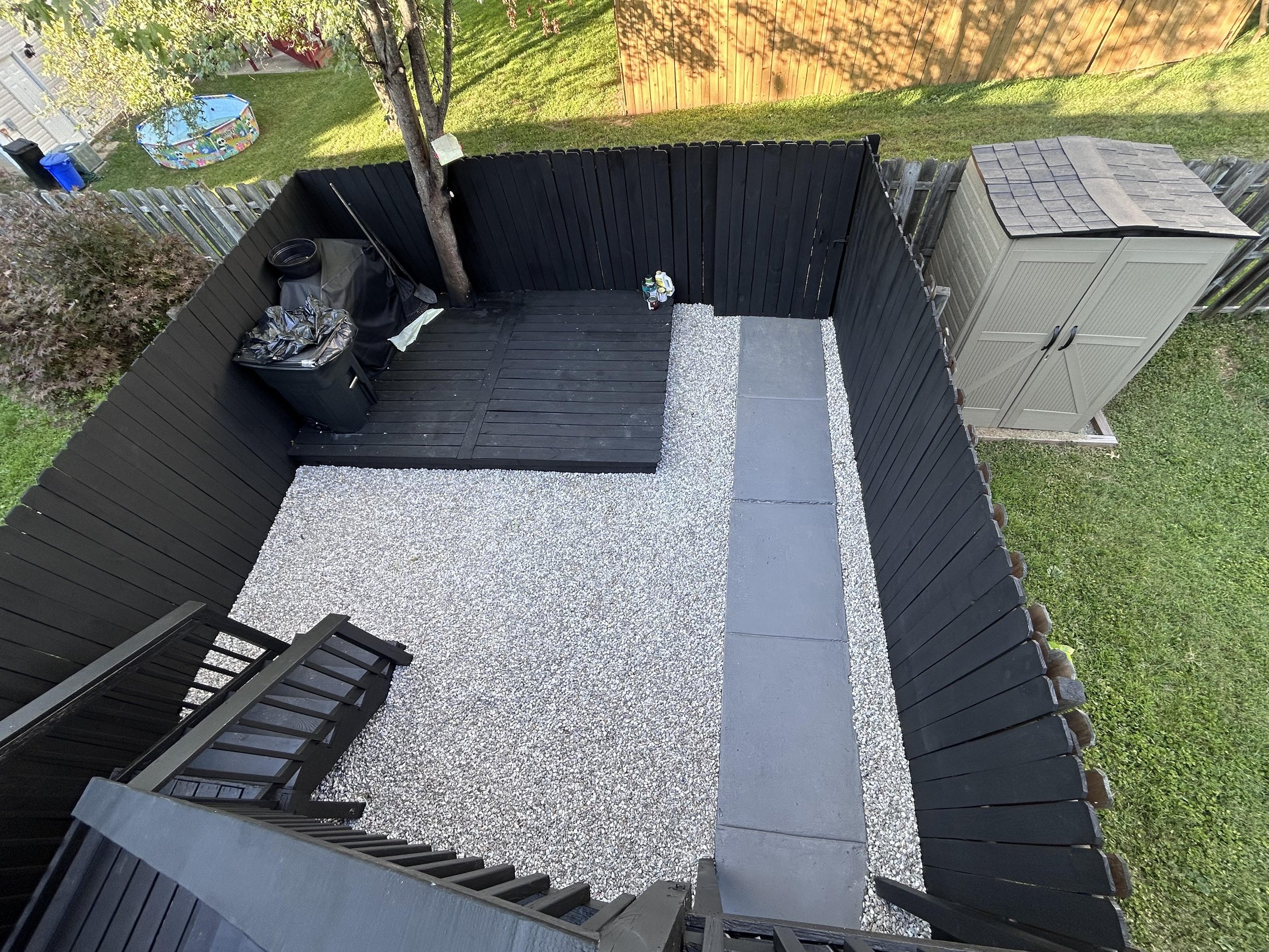 A small backyard patio with black fencing, a gravel area, a concrete path, a storage shed, a tree, and outdoor items including trash bins and a small portable pool.
