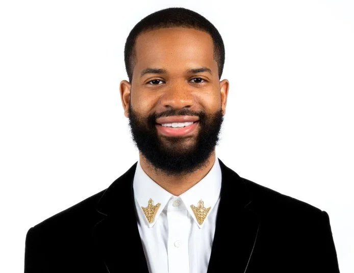 A smiling man with a beard in a black suit with gold embroidered collar details, white shirt, against a white background.