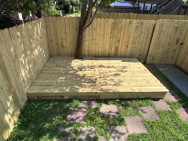 A small outdoor area with a wooden deck, a wooden privacy fence, a tree growing through the deck, and a grassy patch with stone pavers.