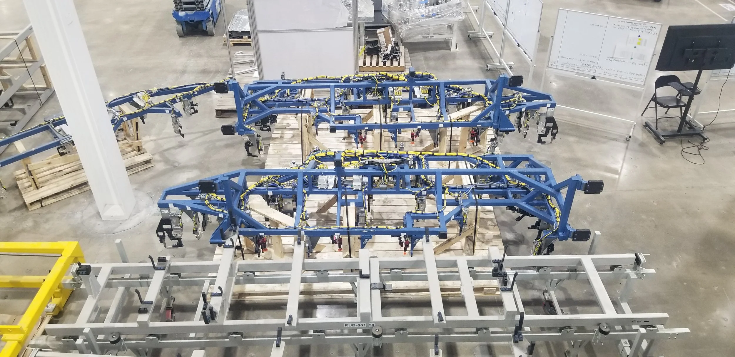 Industrial robotic frames with wiring in a factory, with a whiteboard, a chair, and equipment in the background.