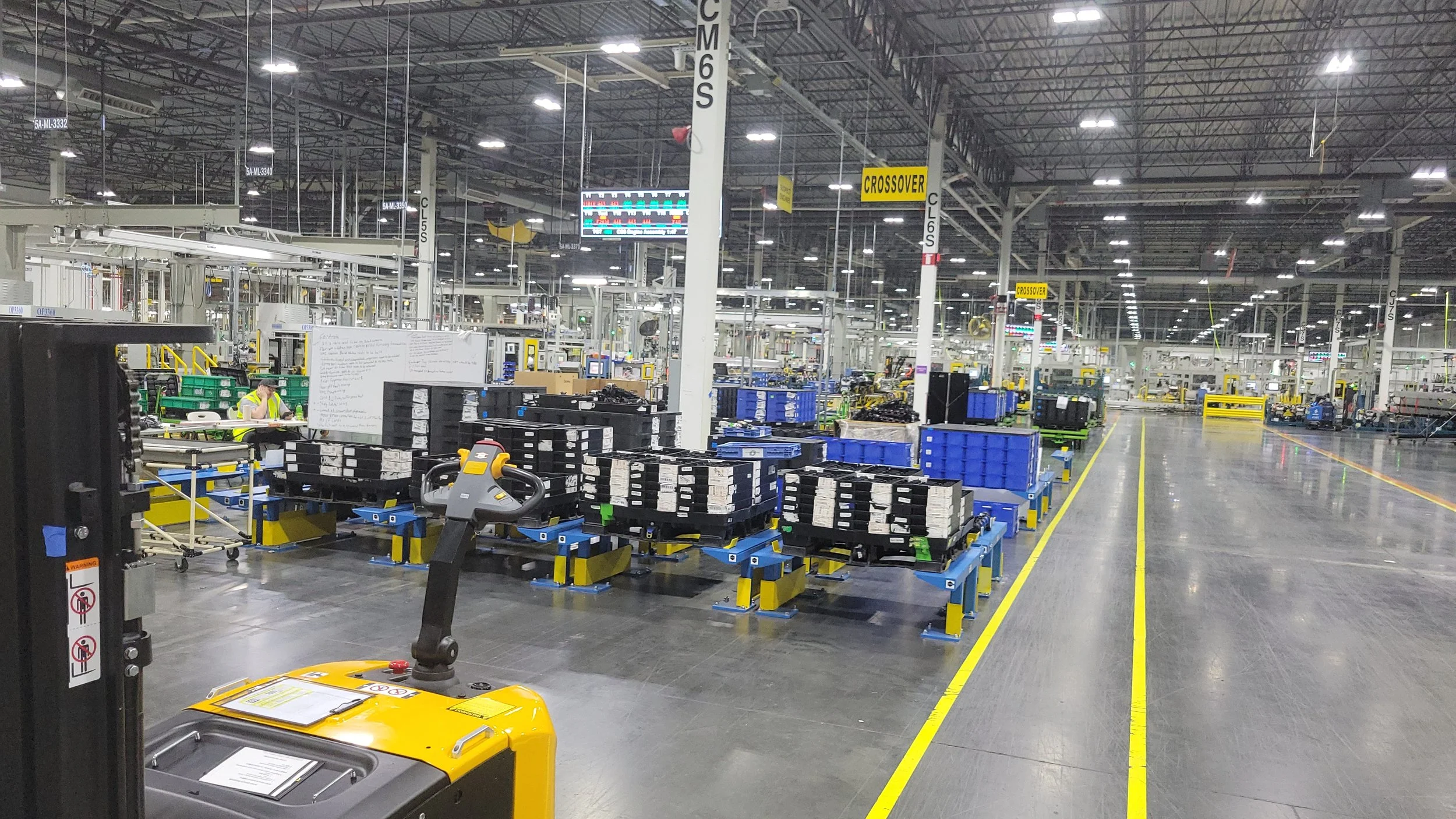 Inside a large, well-lit factory or warehouse with organized work stations, shelves, and equipment, including yellow and black machinery.