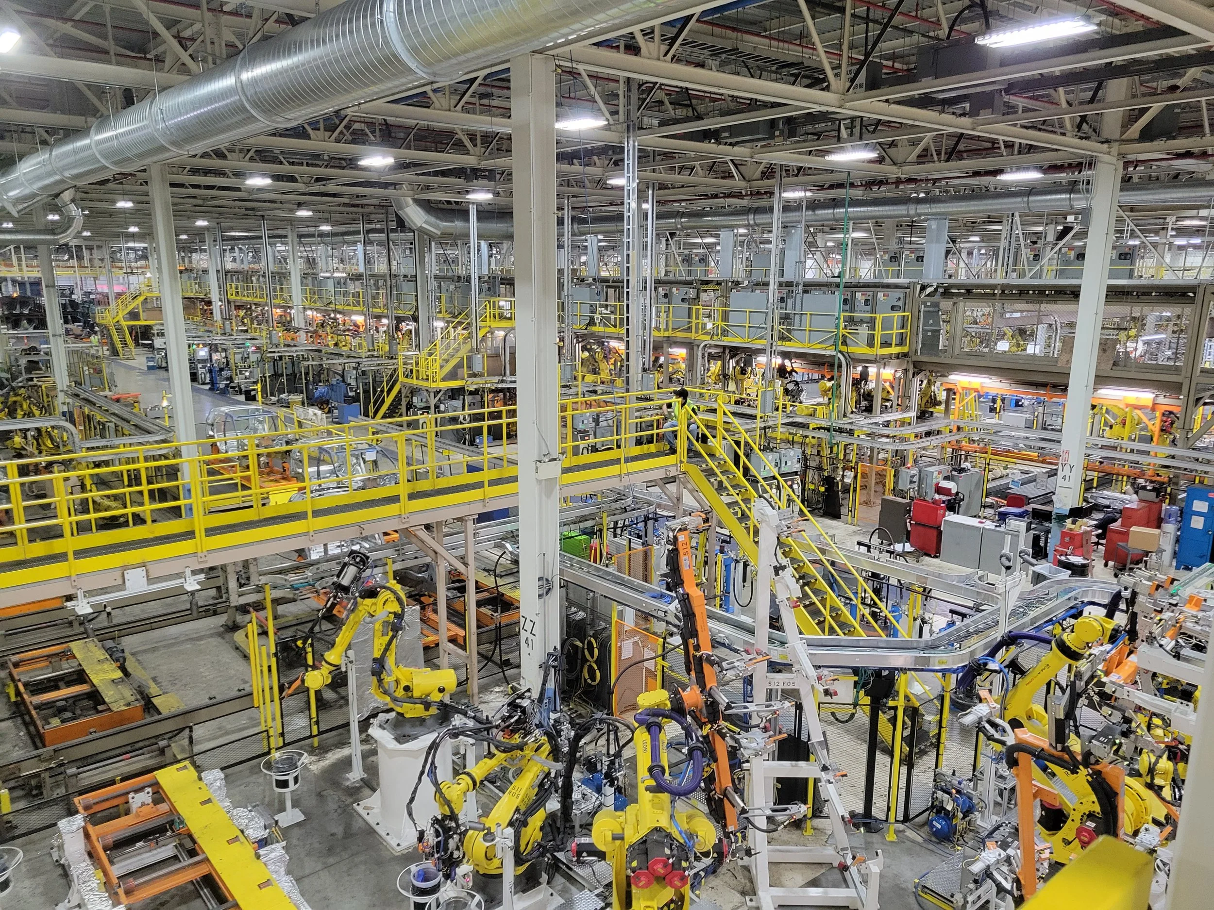 Inside an automotive manufacturing factory with robotic arms, conveyor belts, and yellow safety railings.