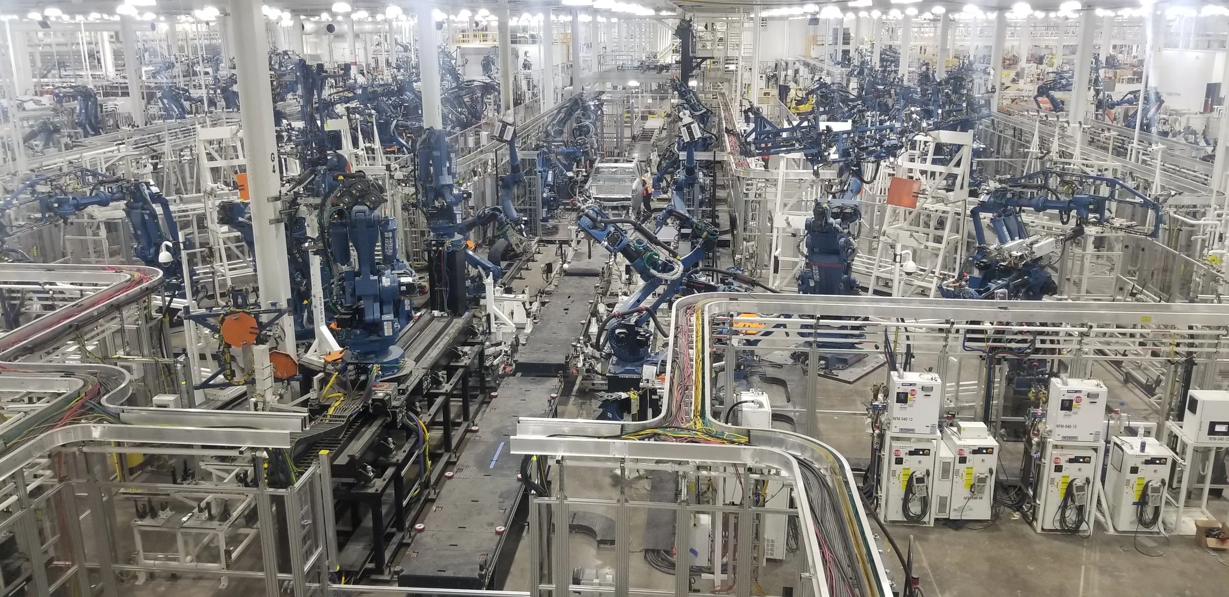 Inside a large factory or warehouse with multiple robotic arms and conveyor systems used for manufacturing or assembly.