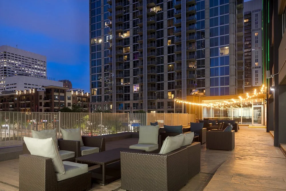 Outdoor rooftop lounge area at night with wicker chairs and cushions, string lights, tall modern buildings in the background, and a view of city streets.