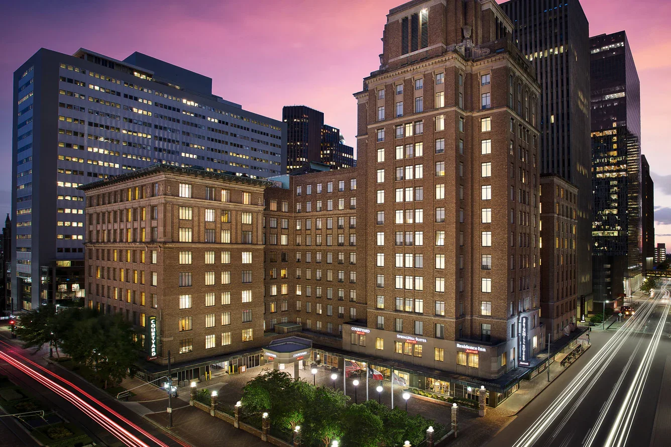Night view of a city street with tall buildings, lights, and traffic trails, featuring a prominent brick hotel building with signs for Courtyard and Residence Inn.