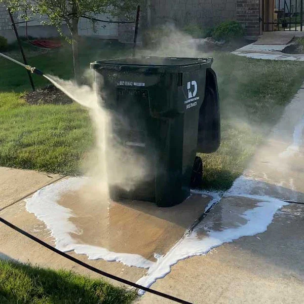 A black trash can on a sidewalk with water spraying from a hose into the can, causing water and soap to overflow onto the sidewalk.
