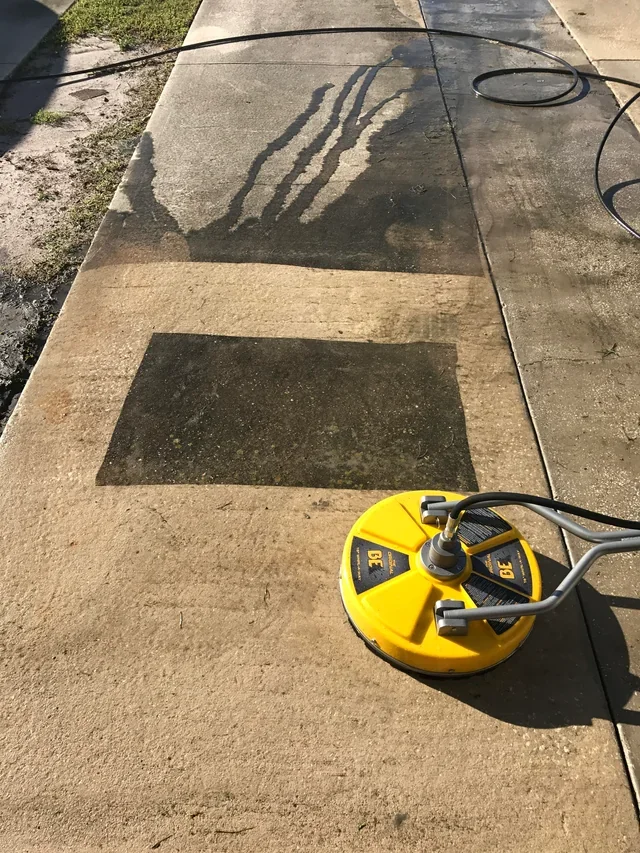 A pressure washer is cleaning a section of concrete sidewalk, creating a pattern of clean and dirty areas with distinct rectangular shapes.