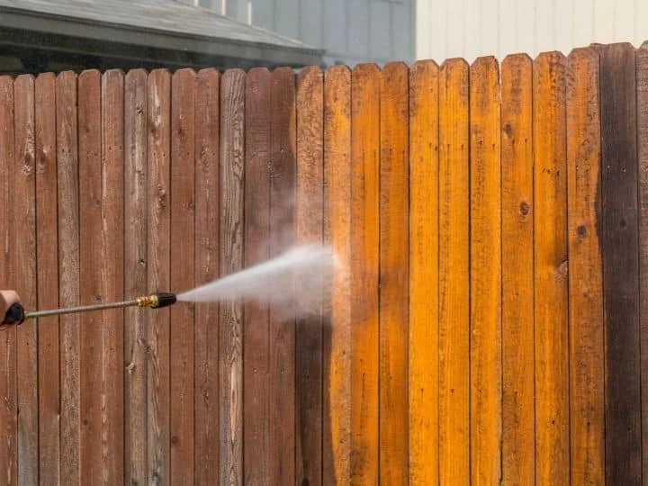 A person using a pressure washer to clean a wooden fence, causing spray and mist.