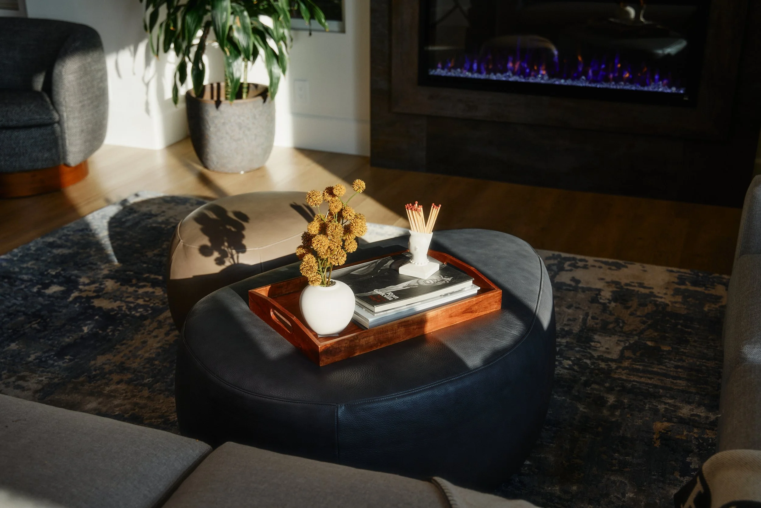 A living room with a gray ottoman and a round coffee table holding a white vase with flowers, a tray, and magazines. In the background, there is a large potted plant and a lit fireplace.