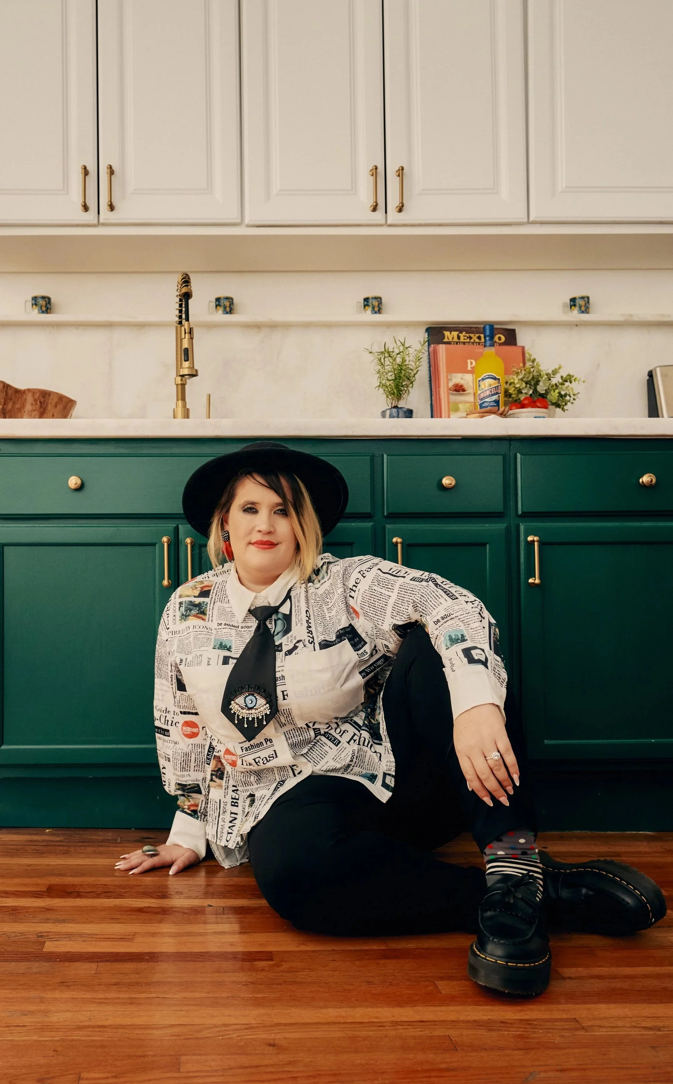 Person wearing a black hat and a newspaper-patterned shirt sitting on the floor in a kitchen with white and green cabinets, hardwood floors, and various decorations and objects on the countertop.