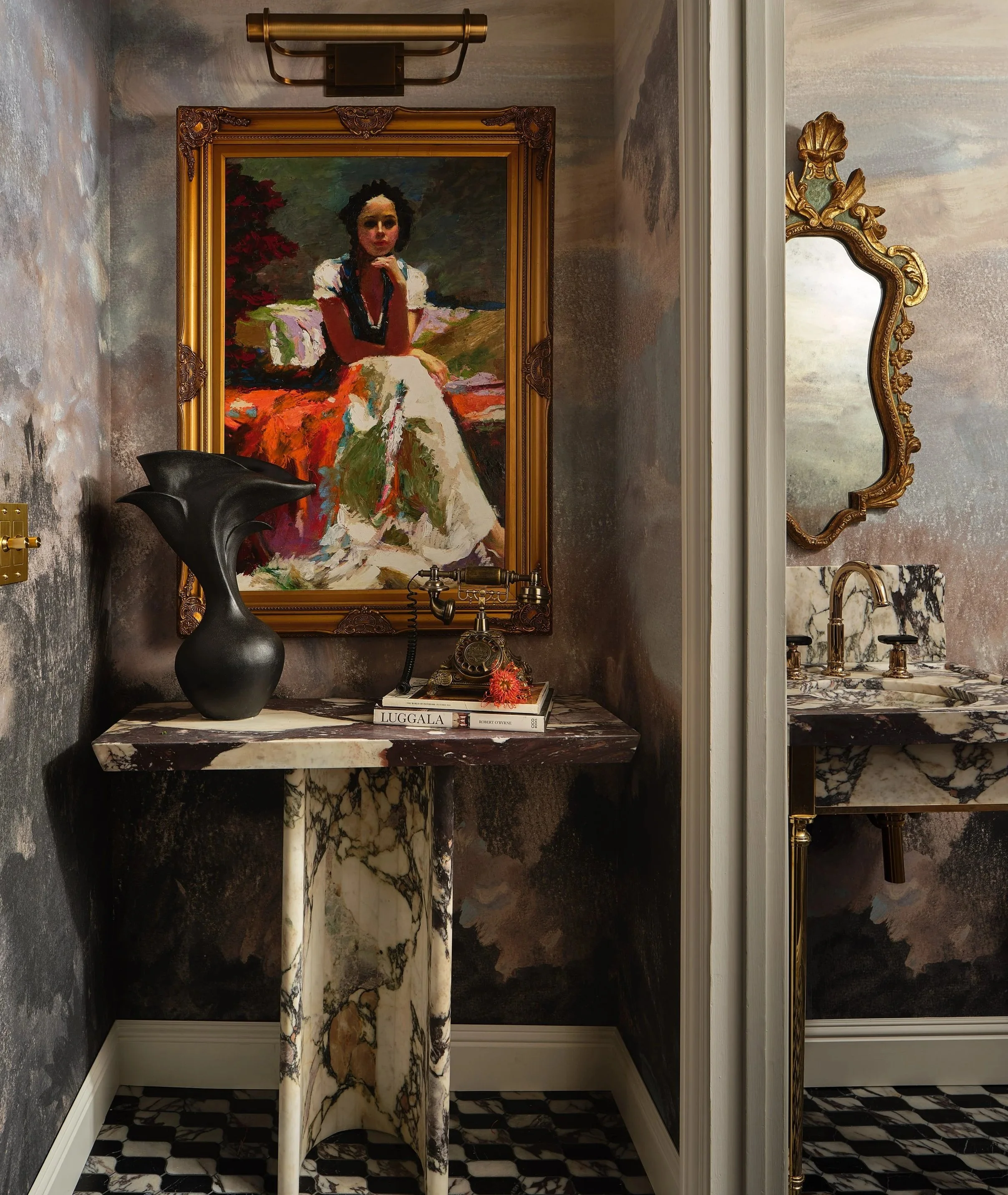 An ornate hallway decor with a framed oil painting of a woman sitting outdoors, a black vase on a marble table, books and a small flower, gold-framed mirror, and a marble countertop and faucet visible on the right.