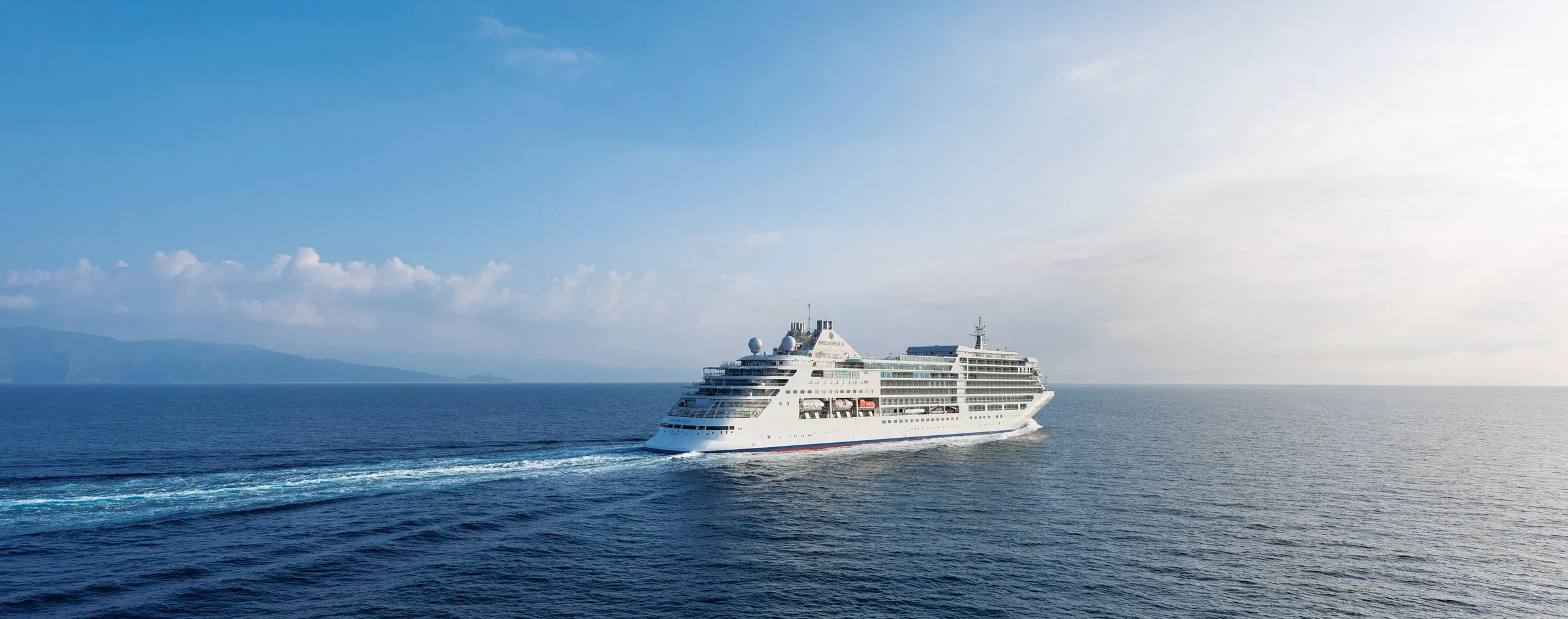 Large Silverseacruise ship sailing on the open ocean with a distant landmass on the horizon, clear sky with some clouds.