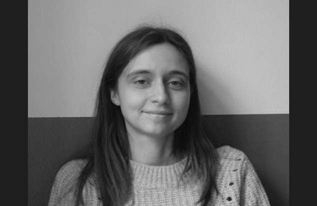 A young woman with long hair and a slight smile, wearing a textured sweater, sitting against a plain wall.
