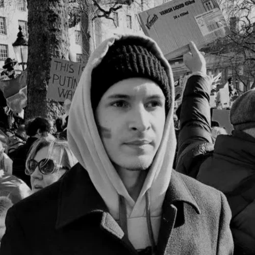 Young man wearing a hoodie and beanie at a protest or rally, with signs and other protesters in the background.