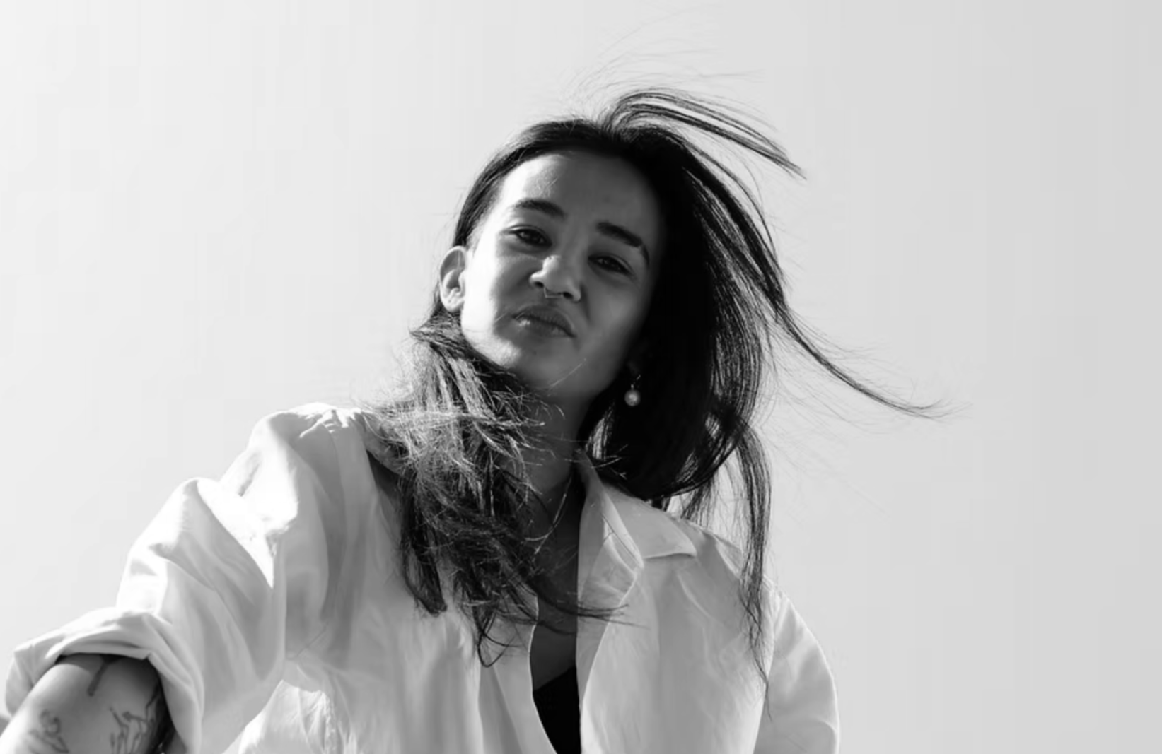 Black and white photo of a woman with long hair, wearing a white shirt with rolled-up sleeves, looking at the camera with a slight smile, hair blowing in the wind.