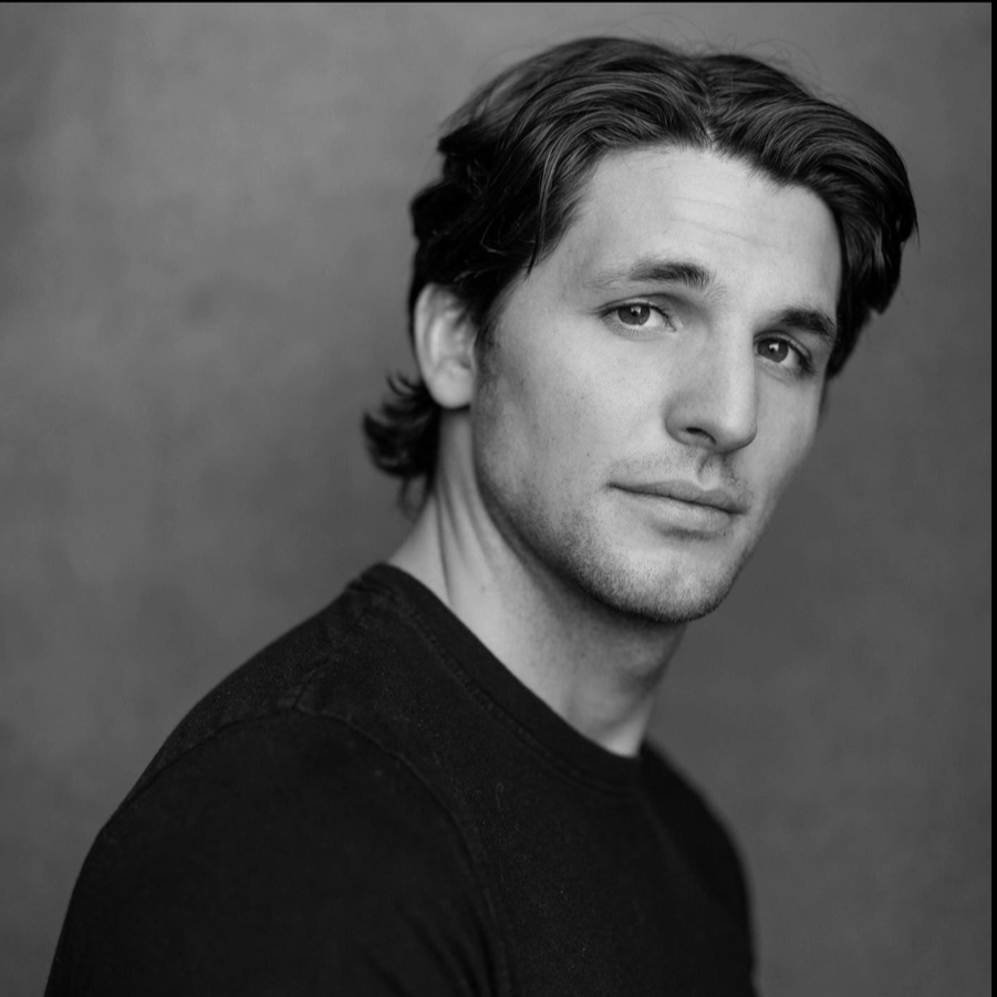 Black and white portrait of a young man with medium-length hair, wearing a dark shirt, looking at the camera with a neutral expression.
