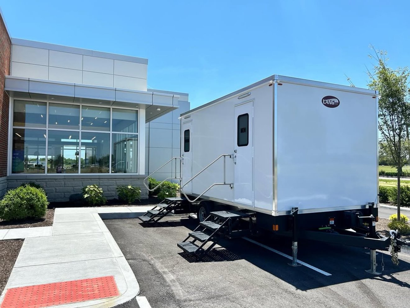 A white mobile restroom trailer with metal steps parked outside a modern building with large glass windows, landscaping, and a sidewalk.