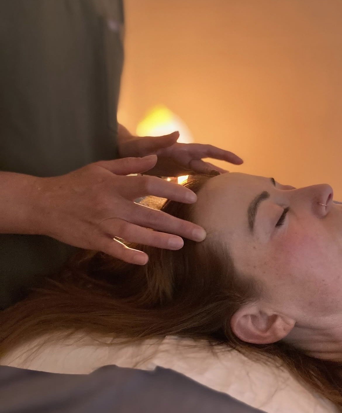 A woman receiving a massage on her head, with hands gently placed on her scalp, in a calming environment with soft lighting.