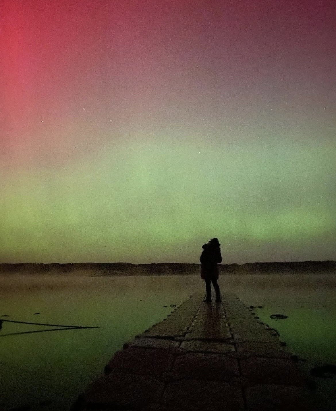 Person standing on a pier looking at the Northern Lights in the night sky.