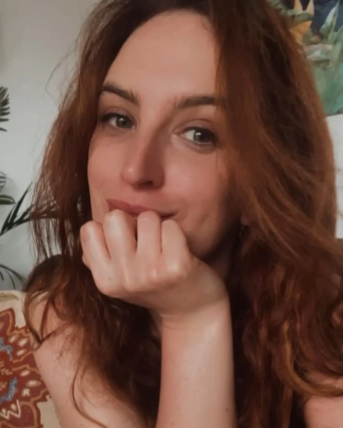 A woman with long wavy red hair resting her chin on her hand, looking at the camera with a slight smile.