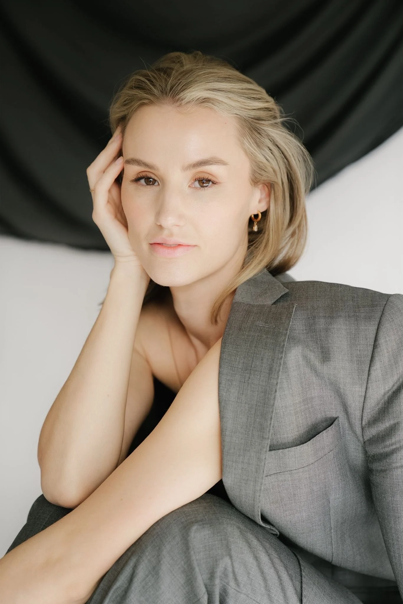 Nuori nainen istuu valkoisessa huoneessa, one-korvikset, harmaa puku, katsookin kameraan.