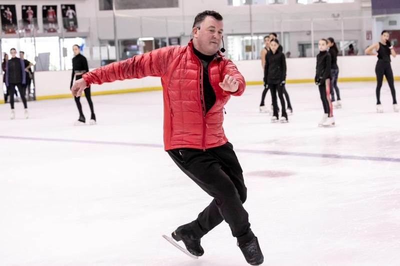 Mies harrastaa taitoluistelua jäällä, muita luistelijoita taustalla, sisäjäähalli.