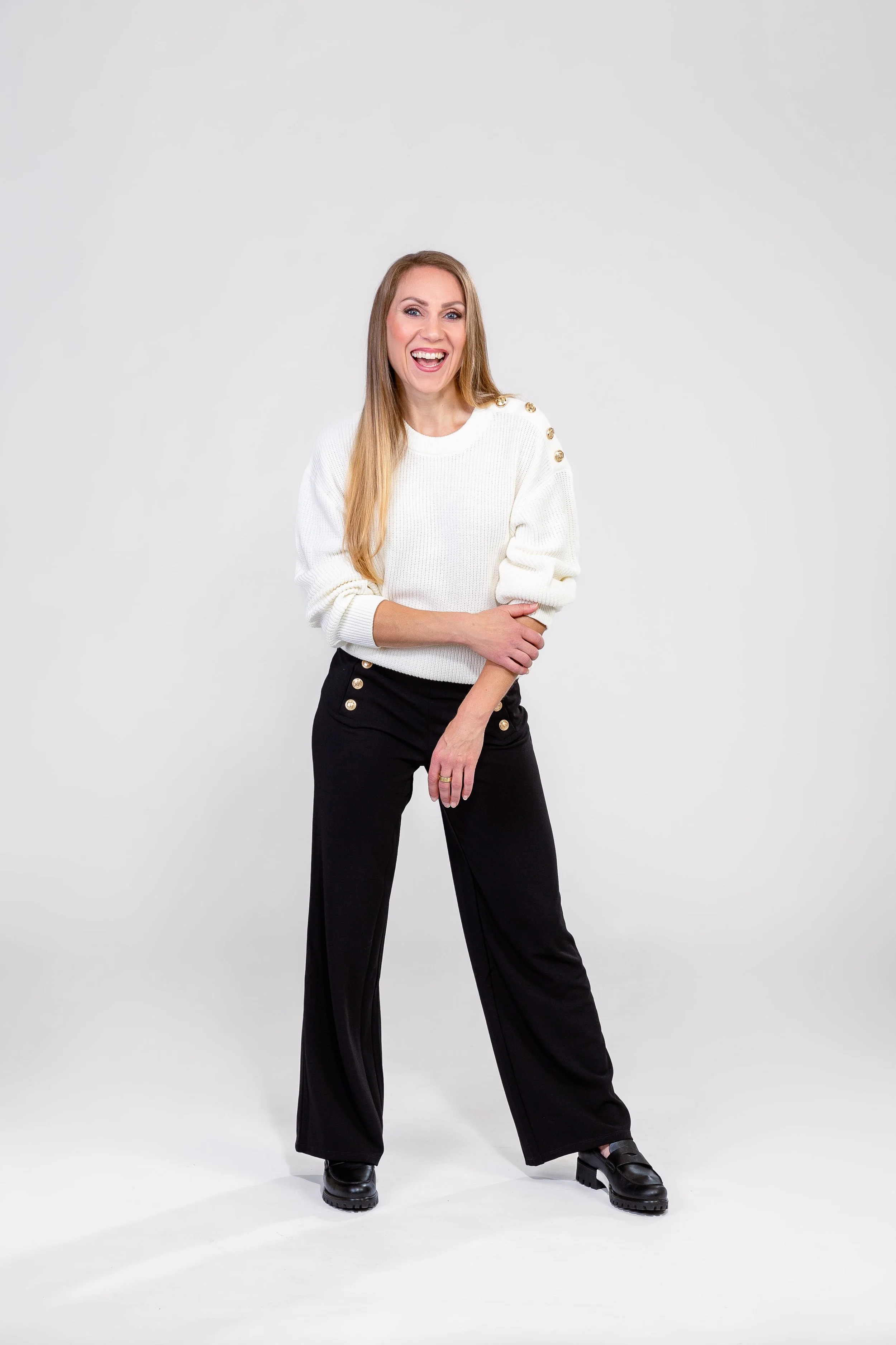 Nainen hymyilee lattialla valkoisessa taustassa, pukeutuneena valkoiseen neuleeseen ja mustiin leveälahkeisiin housuihin, joissa on kullanvärisiä nappeja.