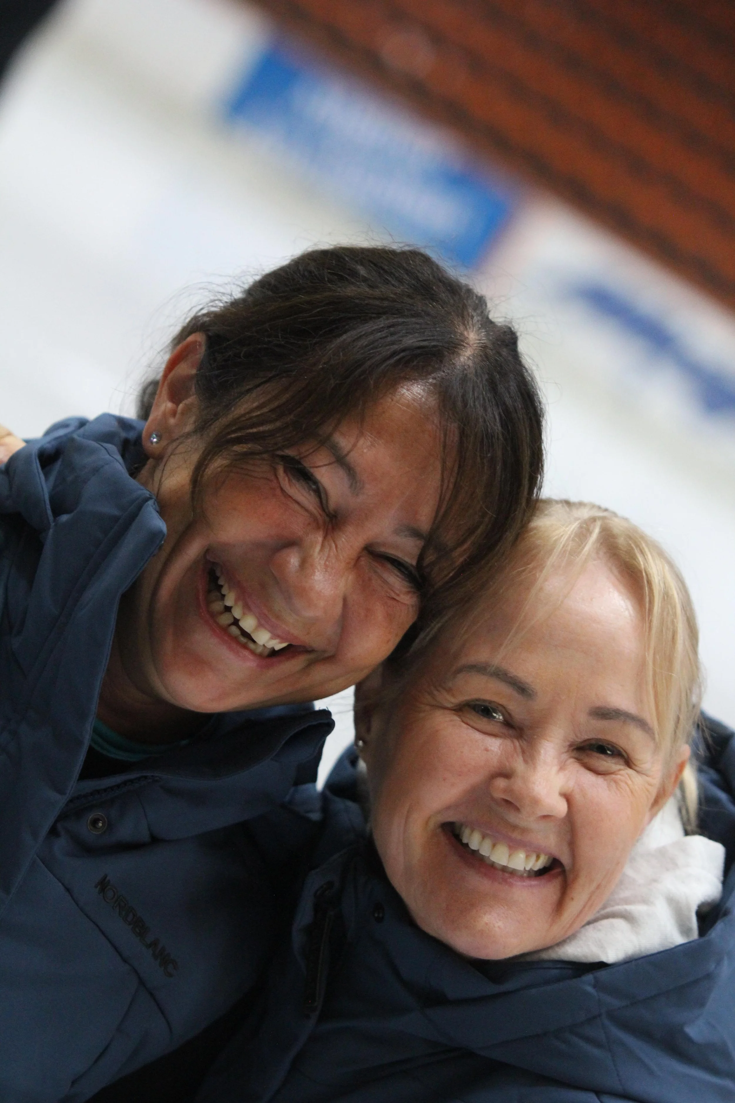 Kaksi naista hymyilevät ja nauravat lähekkäin, pukeutuneina sinisiin ulkoilupukuihin.