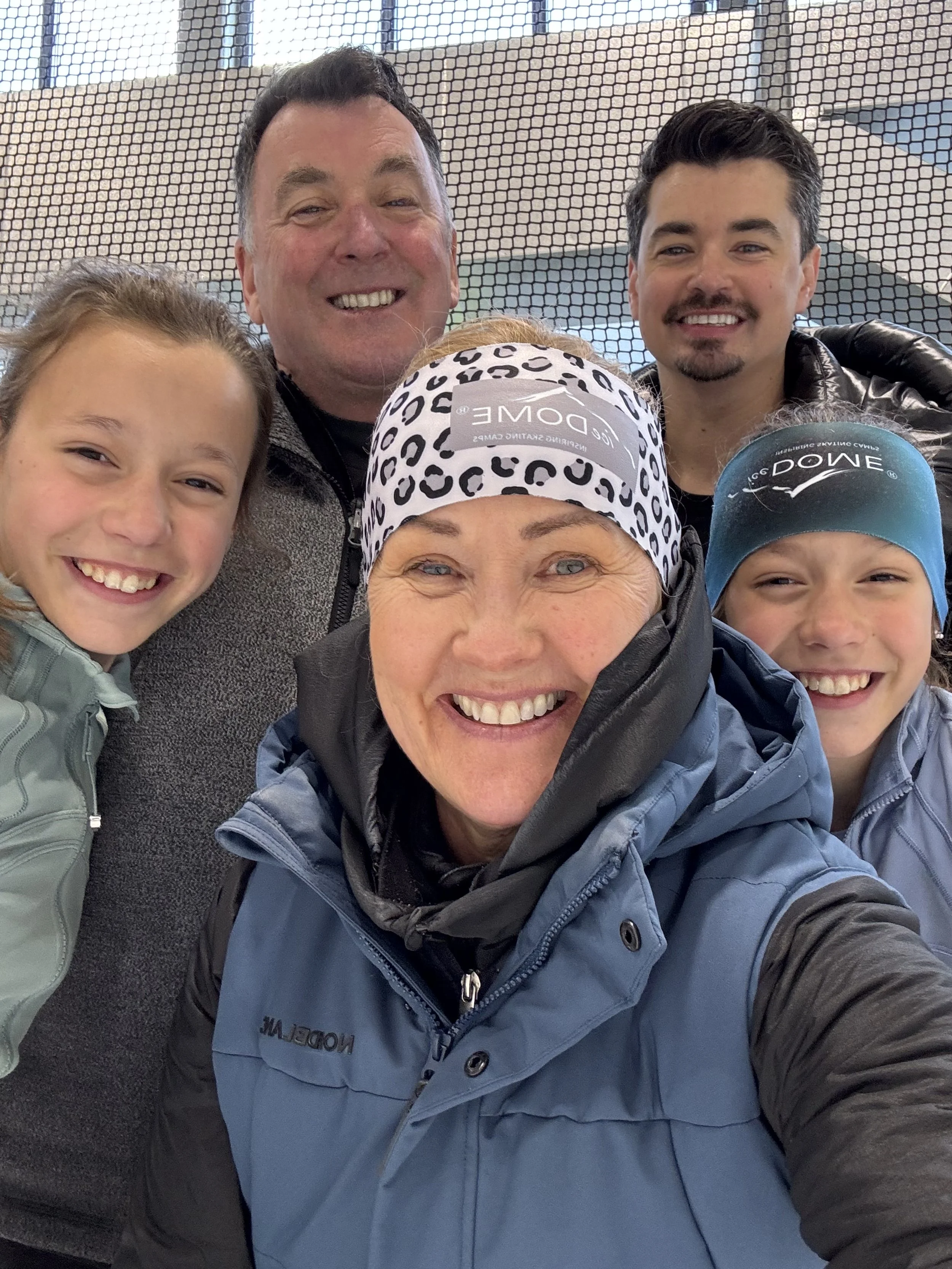 Viisi ihmistä hymyilevät selfiekuvassa. Taustalla pieni verkkoikkuna ja rakennuksia.