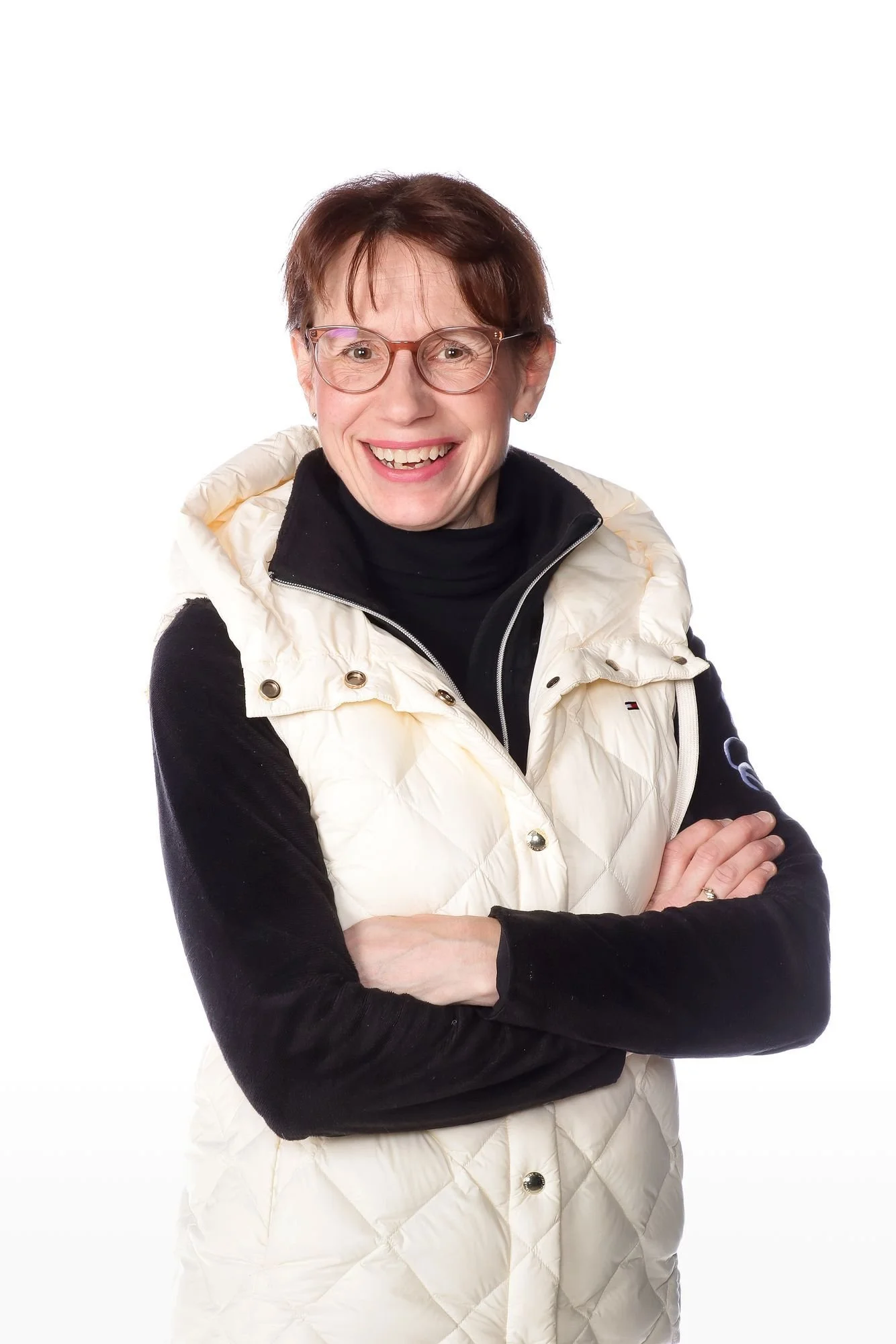 Nainen hymyilee, istuu tai seisoo valkoisella taustalla, päällä ruskettunut hius ja silmälasit, pukeutuneena mustaan pitkähihaiseen ja vaalean beigen untuvatakkiin.