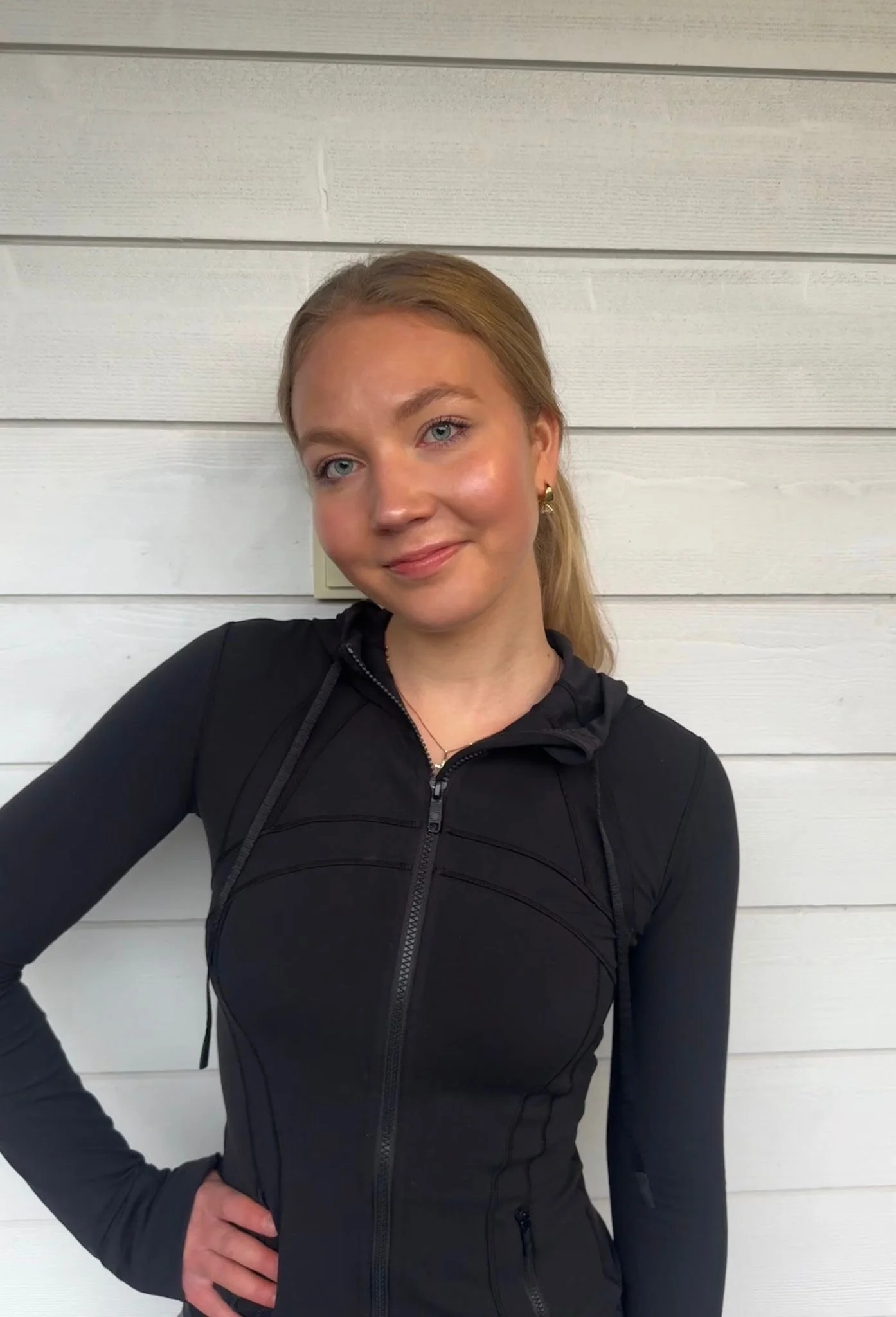 Nuori nainen nojaa seinää vasten asettaen kätensä löysästi lantiolle. Hänellä on vaaleat hiukset ja hän on pukeutunut mustaan urheiluvaatteeseen. Taustalla valkoinen puupaneeli.