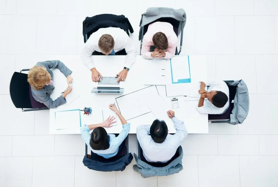 Top-down view of six professionals around a white conference table in a business meeting.