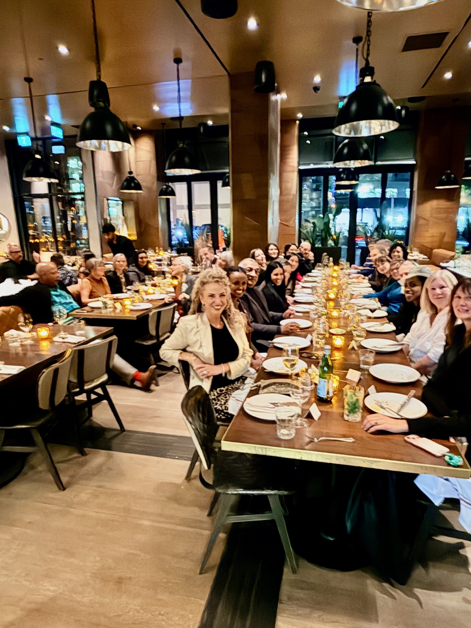 Four women sitting around a table in a cafe, engaging in conversation, with warm orange lighting and brick walls in the background.