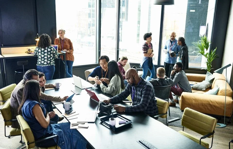 A diverse group of people working and collaborating in a modern office space, some seated at a large table using laptops and tablets, others standing and talking near windows with city views, decorated with a yellow sofa and green plants.