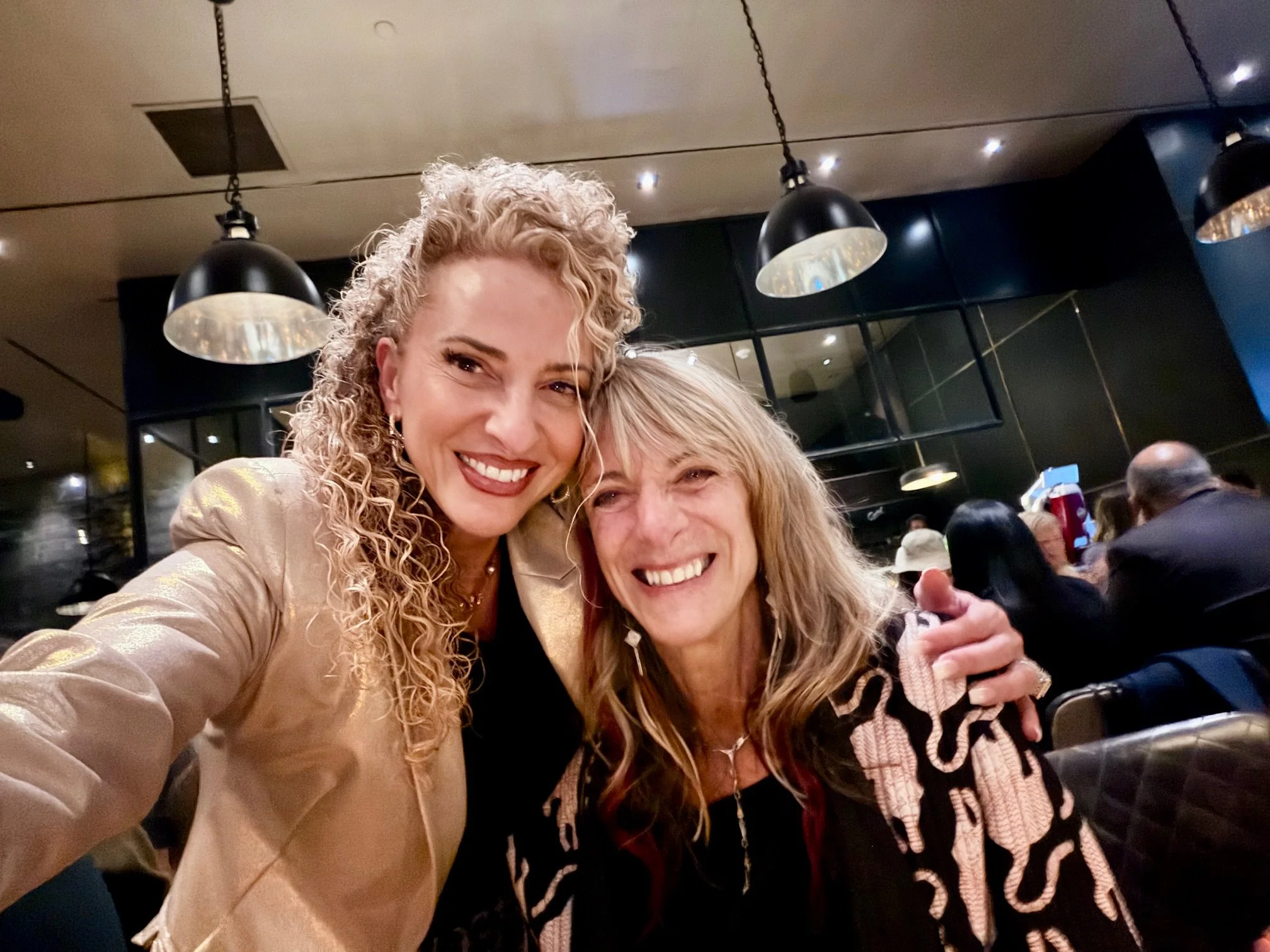 Two women smiling and taking a selfie in a restaurant or cafe, with a dark interior and hanging lights in the background.