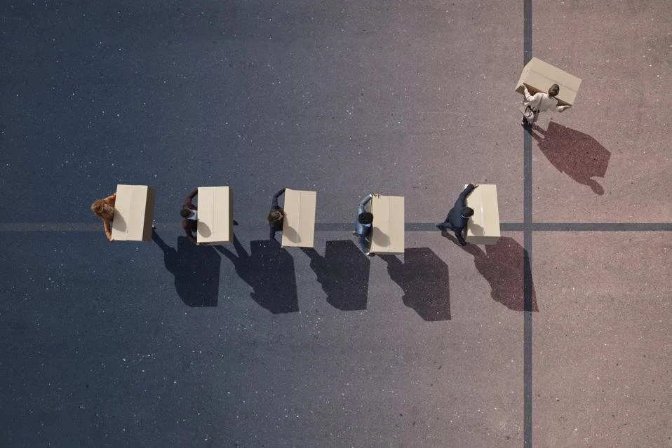 An overhead view of five people standing at separate desks arranged in a line on an outdoor court, with a sixth person pushing a cart with a large box.