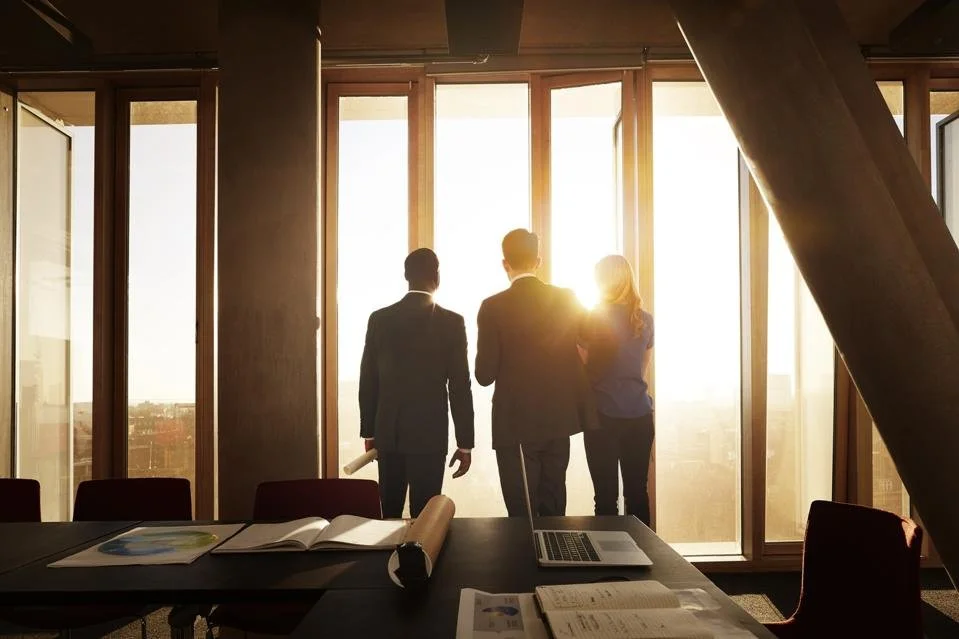 Three business people in suits looking out of large windows as the sun sets, with a conference room table in the foreground.