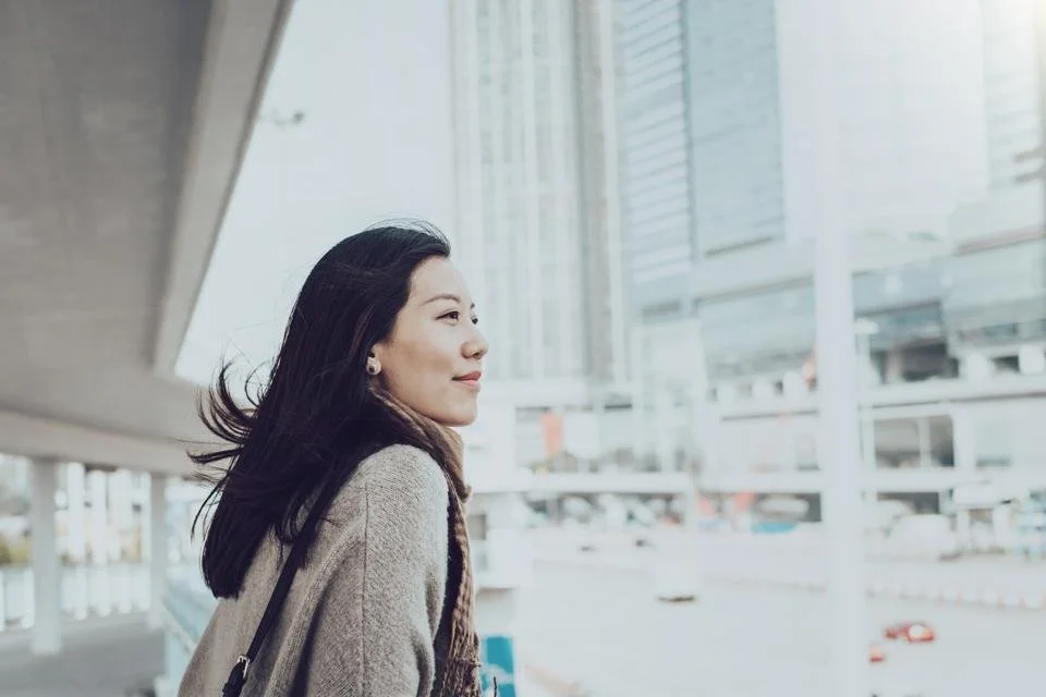 Side view of a woman with long black hair, wearing a beige coat, standing outdoors in an urban area with tall buildings and a parking lot in the background.
